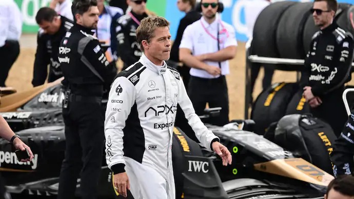Brad Pitt in racing gear beside a Formula 1 car, set against a cheering crowd with box office earnings graph in the background.