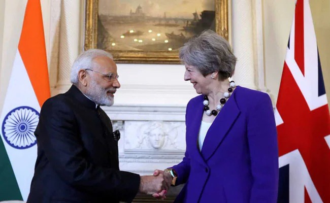 PM Narendra Modi and UK officials shaking hands at a signing ceremony with India–UK flags and FTA documents on the table.
