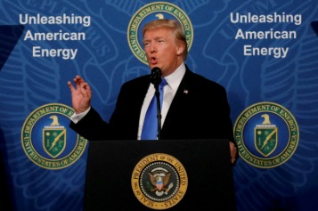 Donald Trump speaking at a press event with oil barrels and world map in background, symbolizing global energy trade.