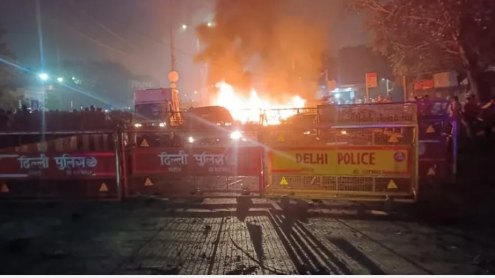 Emergency responders at the site of a car explosion near Delhi’s Red Fort, with smoke rising in the background and police securing the area.