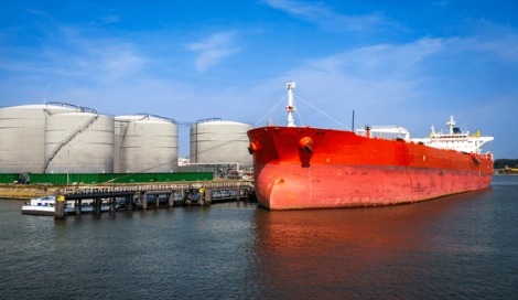 Large crude oil tanker docked at an industrial port with storage tanks in view, representing global oil export and price movement.
